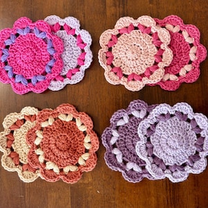May include: A collection of handmade crochet coasters in various colors, including pink, purple, peach, and brown. Each coaster features a circular design with a scalloped edge, perfect for protecting surfaces from drink condensation.