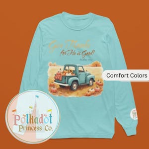 May include: A long-sleeved, light blue shirt with a fall-themed graphic. The graphic features a vintage blue truck filled with pumpkins and sunflowers, with the text "Give Thanks for He is Good!" and "Comfort Colors". A Polkadot Princess Co. logo is on the shirt.