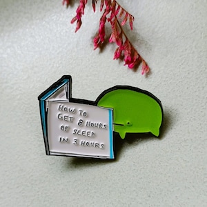 May include: A metal enamel pin featuring a white rectangular book with blue edges and the black text "HOW TO GET 8 HOURS OF SLEEP IN 3 HOURS" next to a rounded, bright green creature with a small black eye and two short legs on the right side.