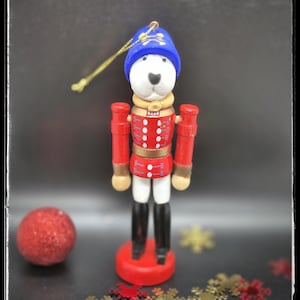 May include: A wooden Christmas ornament shaped like a dog in a red uniform and blue hat. The dog has a white face and black boots. The ornament is on a red base and has a gold string for hanging. The text "HANDMADE BULLTERRIERMANIASHOP" is at the bottom.