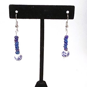 Può includere: Un paio di orecchini pendenti con perline blu e viola e un ciondolo in ceramica bianca con motivi floreali blu. Gli orecchini sono esposti su un espositore per orecchini nero.