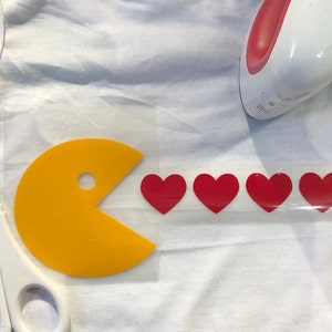 May include: A yellow Pac-Man character with an open mouth, eating four red hearts. The image is on a clear sheet of transfer paper, ready to be ironed onto a fabric surface.