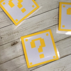 May include: Square white stickers with a yellow border and a yellow question mark design. The stickers have a pixelated design, reminiscent of a video game. Several stickers are visible on a light wood surface.