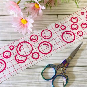 May include: A sheet of clear adhesive vinyl with a grid pattern and pink bubble shapes cut out. A pair of rainbow-colored scissors is lying on a wooden surface.