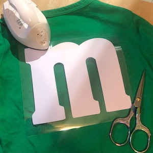 May include: A white vinyl letter 'M' on a clear sheet, ready to be ironed onto a green t-shirt. A white iron and silver scissors are also in the image.