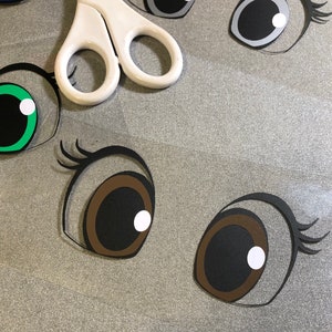 May include: A close-up of several paper craft eyes, including brown, black, green, blue, and grey eyes. The eyes are cut out and ready to be used for a craft project.
