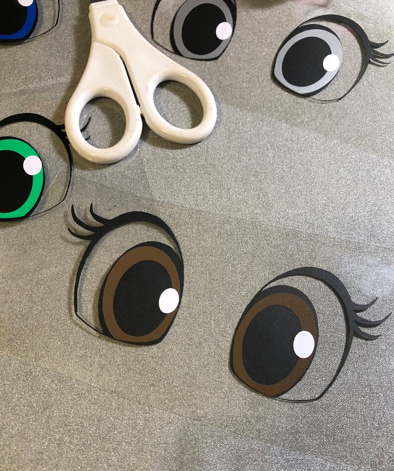 May include: A close-up of several paper craft eyes, including brown, black, green, blue, and grey eyes. The eyes are cut out and ready to be used for a craft project.