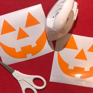 May include: Two square sheets of white backing paper featuring orange vinyl jack-o'-lantern face decals with two triangular eyes, a triangular nose, and a wide grinning mouth. A white handheld craft iron and white scissors are positioned nearby.