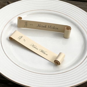 May include: Two gold scroll place cards on a white plate. The top card reads "Table 1 Niamh Mahon" and the bottom card reads "Table 2 Kevin Wilson".