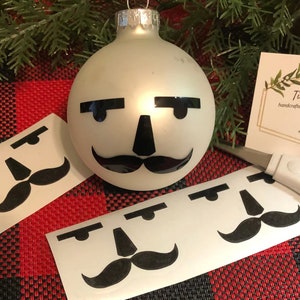 May include: White Christmas ornament with a black mustache, eyebrows, and nose.  The ornament is surrounded by sheets of white paper with black mustache, eyebrows, and nose stickers.