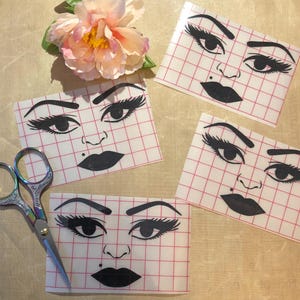 May include: Four face decals with a black and white design, featuring bold eyebrows, long eyelashes, and a beauty mark. A pair of decorative scissors and a pink and yellow flower are also in the image.