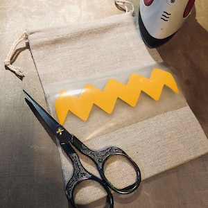 May include: A close-up of a beige fabric bag with a yellow zig-zag design on a clear plastic sheet. A pair of black scissors with gold accents are lying on the bag. An iron is visible in the background.