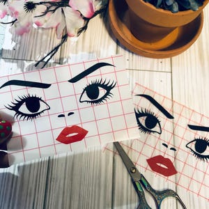 May include: Two white sheets of paper with a grid pattern. Each sheet has a black and white illustration of a woman's face with red lips. The face on the top sheet has both eyes and eyebrows. The face on the bottom sheet has one eye and eyebrow. A pair of rainbow-colored scissors is on the table.