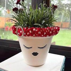 May include: A white ceramic plant pot featuring a black graphic of closed eyes with long eyelashes and dark lips. The rim is adorned with a red fabric cover featuring white polka dots and a circular rhinestone embellishment holding a green plant.