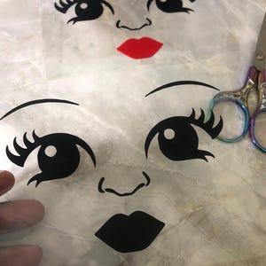 May include: Two transparent sheets with black facial features, including eyes, eyebrows, noses, and lips. One sheet has a red lip. A pair of scissors with rainbow handles is in the background.