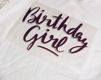 May include: A white V-neck t-shirt featuring a dark maroon graphic transfer on the front. The graphic consists of the words Birthday Girl written in a cursive script font. The transfer is applied to the center of the shirt chest area.