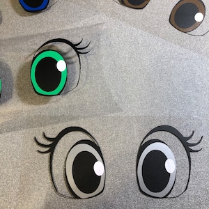 May include: A set of six large googly eyes with black pupils and white highlights. The eyes are made of paper and have different coloured rims, including blue, green, brown, and silver.