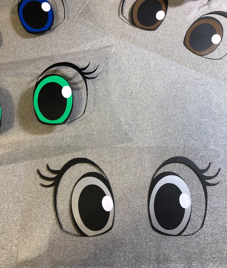 May include: A set of six large googly eyes with black pupils and white highlights. The eyes are made of paper and have different coloured rims, including blue, green, brown, and silver.