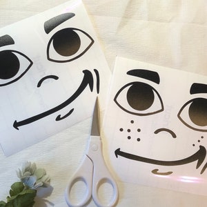 May include: Two white sheets of paper with black ink drawings of cartoon faces. The faces have large eyes, eyebrows, and a smiling mouth. The faces are on a white surface.