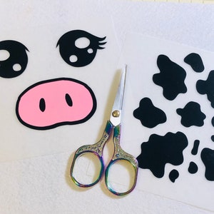 May include: Two sheets of white vinyl with black and pink cutouts. One sheet has a cow face with eyelashes, a pink nose, and two black eyes. The other sheet has black cow spots. A pair of rainbow-colored scissors is in the middle of the sheets.