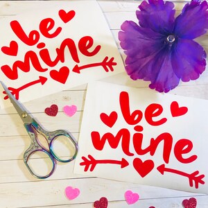 May include: Two white sheets of paper with the phrase "be mine" written in red cursive letters with red hearts and arrows. The sheets are on a white wooden surface with pink and red glitter hearts scattered around.