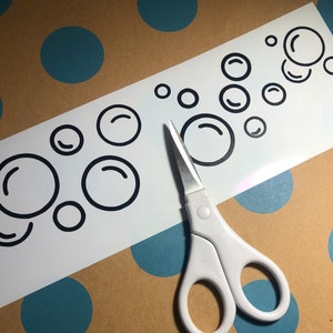 May include: A white sheet with a black bubble design and a pair of white scissors. The scissors are open and positioned near the bubbles. The background is a brown surface with blue polka dots.