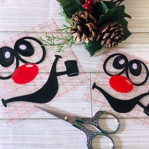 May include: Two black and red cartoon face decals with a red nose, black eyes, and a black mustache holding a black mug. A pair of decorative scissors and holiday greenery with pinecones are also in the image.