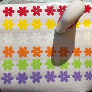May include: A sheet of white paper with rows of colorful flower stickers. The stickers are in various colors, including red, yellow, white, orange, green, and purple. A heat press is visible in the upper right corner of the image.