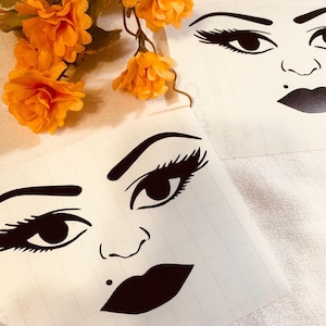 May include: Two black and white illustrations of a woman's face with dramatic eyelashes, bold eyebrows, and a full, dark lip. The illustrations are printed on white paper.