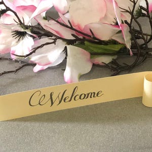 May include: A yellow paper scroll with the word "Welcome" written in black cursive script. The scroll is rolled up at both ends and is lying on a gray surface. There are pink and white flowers in the background.