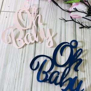May include: Two decorative "Oh Baby" signs, one in pink and one in blue. The signs are made of paper and have a cursive font.