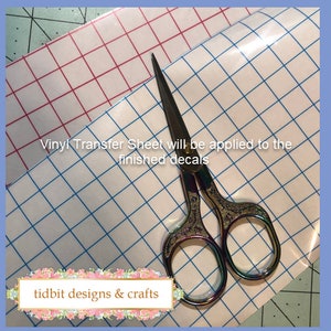 May include: A clear vinyl transfer sheet with a blue grid pattern. A pair of rainbow-coloured scissors is on top of the sheet. The text "Vinyl Transfer Sheet will be applied to the finished decals" is visible on the sheet. The text "tidbits designs & crafts" is visible in the bottom left corner of the image.