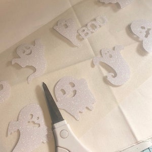 May include: A white sheet of paper with several white glitter ghost cutouts and the word "BOO" on it. A pair of white scissors is on the paper.