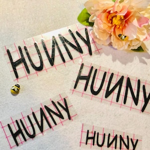 May include: Four sheets of clear vinyl with the word "Hunny" written in black script on each sheet. The sheets are on a white surface with a pink grid pattern.