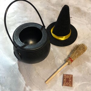 May include: A black plastic cauldron with a handle, a black felt witch hat with a yellow ribbon band, a small straw broom with a red band, and a miniature rectangular book with a brown and gold decorative cover design.