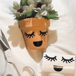 May include: A terracotta flower pot with a smiling face and eyelashes. The pot is decorated with black vinyl stickers. The pot is filled with a small arrangement of artificial flowers.
