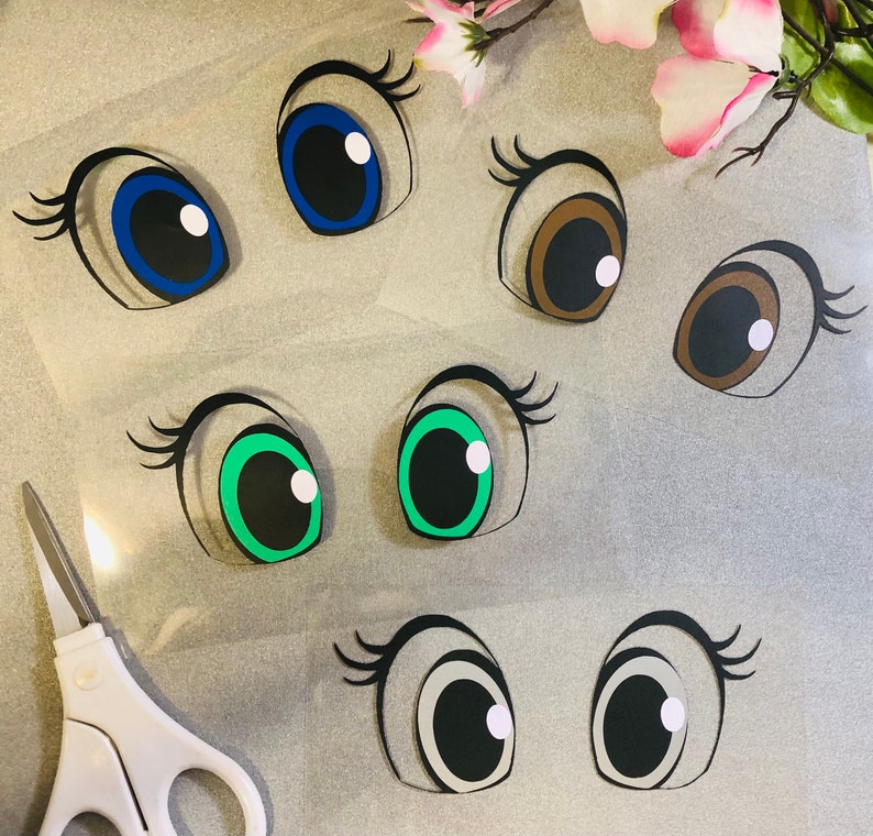 May include: Six pairs of cartoon eyes cut out of paper. The eyes are in various colours, including blue, brown, green, and grey.
