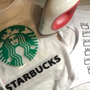 May include: A white t-shirt with a green Starbucks logo and the text "STARBUCKS" in black. A heat press is on top of a white t-shirt with a list of coffee options: Decaf, Shots, Syrup, Milk, Custom, Drink.
