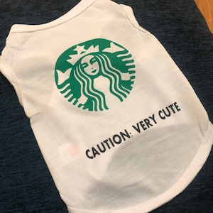 May include: White sleeveless dog shirt with a green Starbucks logo and the text "CAUTION: VERY CUTE" printed on the front.