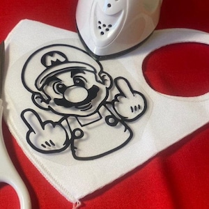 May include: A white fabric face mask with a black and white iron-on design of a video game character giving the middle finger. The design is on a clear sheet of transfer paper.