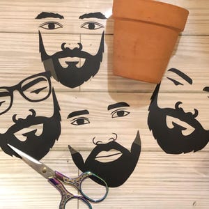 May include: Four black and white paper cutouts of faces with beards. The faces are arranged on a wooden surface. A pair of scissors is in the foreground.