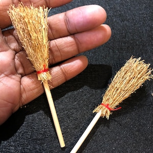 May include: Two miniature brooms with brown straw bristles and red string tied around the base of the bristles. The brooms have wooden handles.