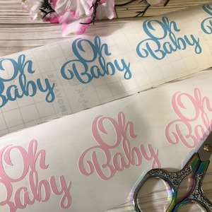 May include: Two sheets of white vinyl with the phrase "Oh Baby" written in cursive. The top sheet has the phrase in light blue, and the bottom sheet has the phrase in pink. A pair of silver scissors is visible in the bottom right corner.