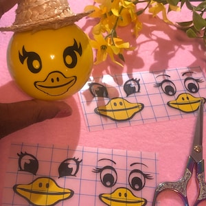 May include: A yellow rubber ball with a duck face and a straw hat. The ball is being decorated with stickers of duck eyes, eyelashes, and beaks. The stickers are on a sheet with a grid pattern.