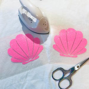 May include: Two pink glitter seashell iron-on transfers on white paper with a small white iron and silver scissors.
