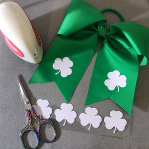 May include: A green cheer bow with white shamrock designs. There are also four white shamrock stickers on a clear sheet. A pair of scissors and a heat press are also in the image.