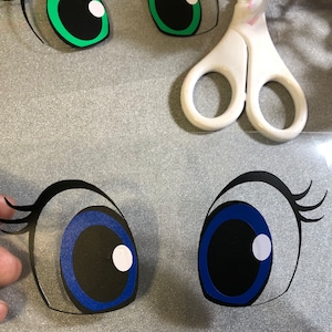 May include: Two pairs of cartoon eyes, one pair green with black pupils and the other pair blue with black pupils. The eyes are cut out of paper and are ready to be used for a craft project.