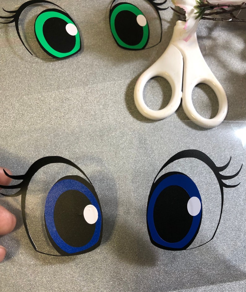 May include: Two pairs of cartoon eyes, one pair green with black pupils and the other pair blue with black pupils. The eyes are cut out of paper and are ready to be used for a craft project.