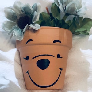 May include: A terracotta flower pot painted with a Winnie the Pooh face. The pot has black eyes, a black nose, and a black smiling mouth. The pot is filled with artificial green plants and white flowers.