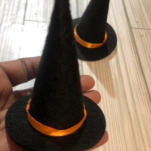 May include: Two miniature black witch hats with orange ribbon bands. The hats have a conical shape and a circular brim. The hats are made of a felt-like material. The image is taken from a close-up angle.